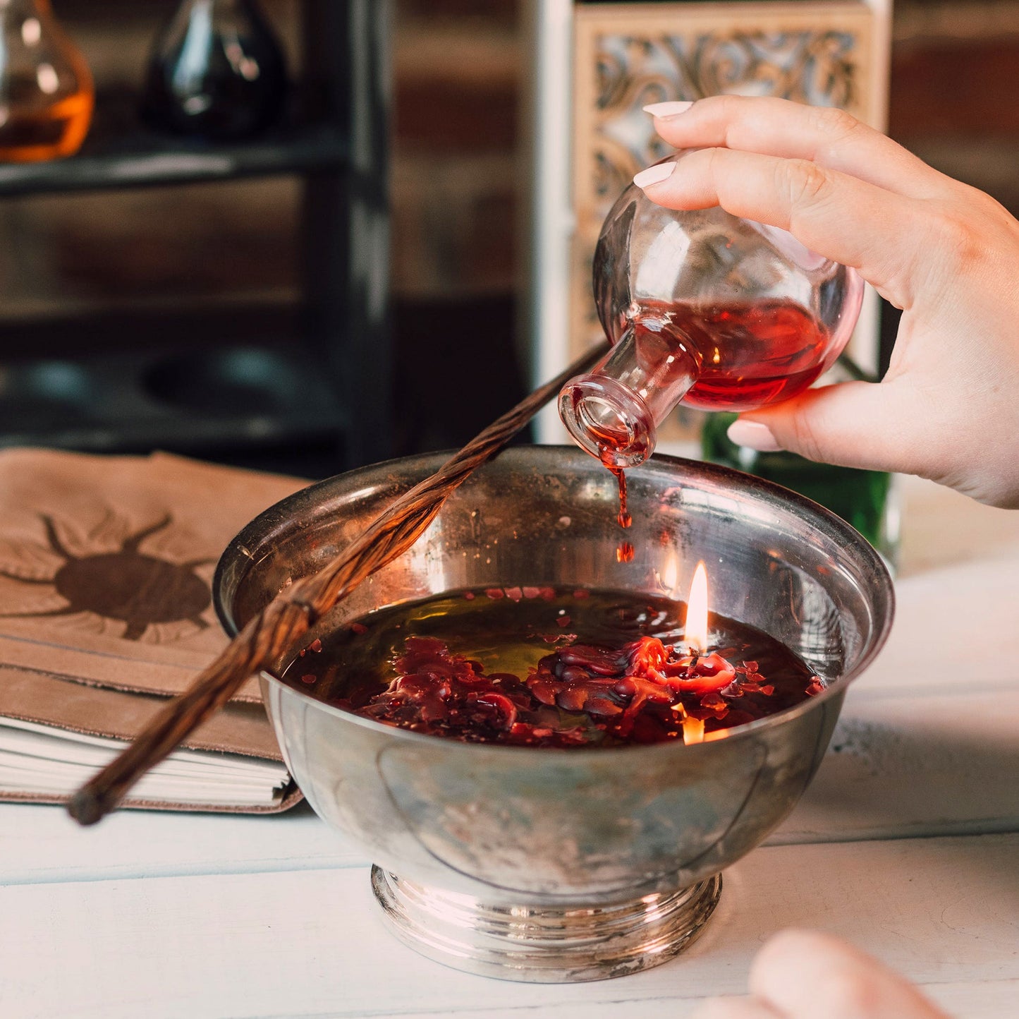 Hand pouring red liquid from a small glass container into a larger metal bowl on a table.